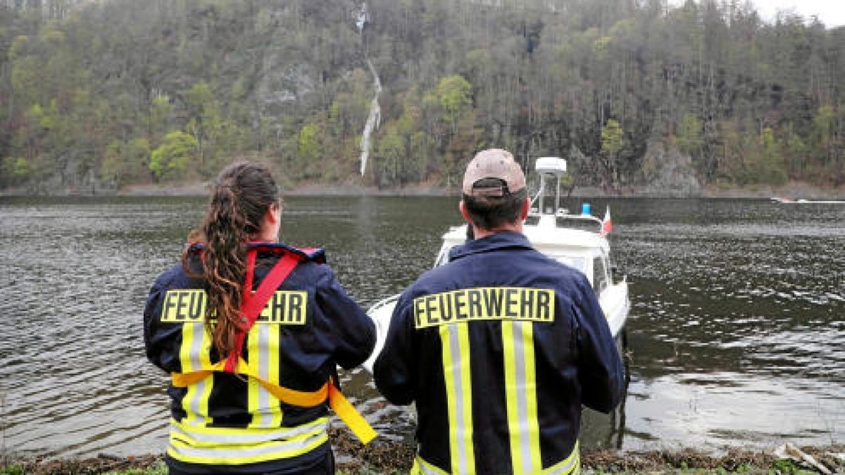 Feuerwehrleute stehen bei einem Brand im Wald an der Bleilochtalsperre am Rande des Wassers und blicken auf das gegenüberliegende Ufer. Foto: Bodo Schackow/dpa