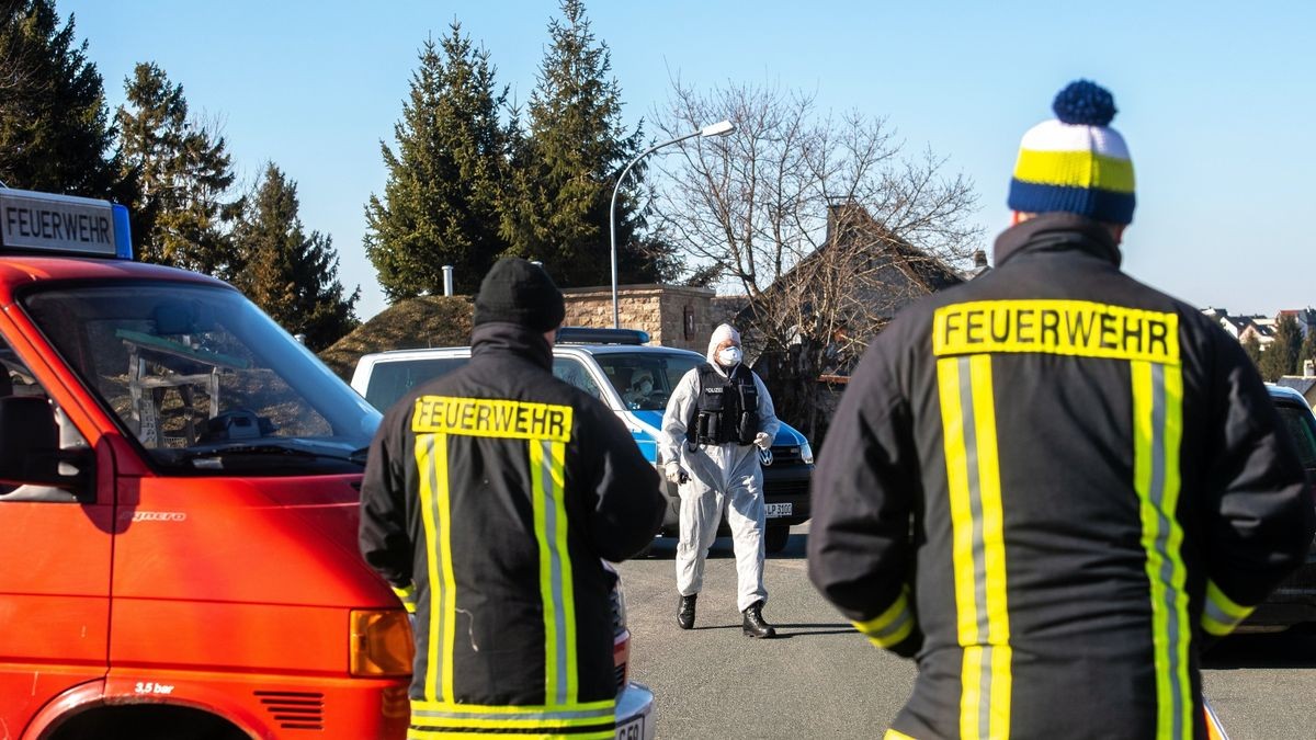 Ein Polizist in Vollschutzanzug sowie Feuerwehrleute stehen am Ortseingang von Neustadt am Rennsteig.