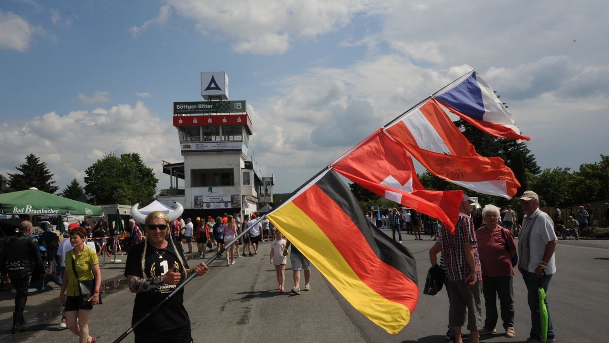 Zdenek Sedlak aus Brünn mit einem Fahnenmast mit deutscher, schweizerischer, österreichischer und tschechischer Flagge. Der überwiegende Teil der rund 600 Fahrzeuge auf zwei, drei und vier Rädern, die an der Veranstaltung „Schleizer Dreieck 100“ anlässlich des Rennstreckenjubiläums teilnahmen, beteiligte sich am Samstagnachmittag bei der Nachstellung des historischen Starts von 1923. Über 70 Komparsen in Bekleidung der ersten Jahrzehnte des 100 Jahre alt gewordenen Schleizer Dreiecks standen nahe des Gasthauses Weidmannsruhe bei Schleiz-Oberböhmsdorf am Rand der Rennstrecke. Sie verfolgten wie Stadtarchivar Ingo Möckel den damaligen Rennleiter Johannes Wächter spielte und die Strecke freigab. Heimatgeschichtlicher Juergen K. Klimpke verkörperte den Schleizer-Dreieck-Entdecker Karl Slevogt, der in einem Apollo-Fahrzeug mitfuhr.