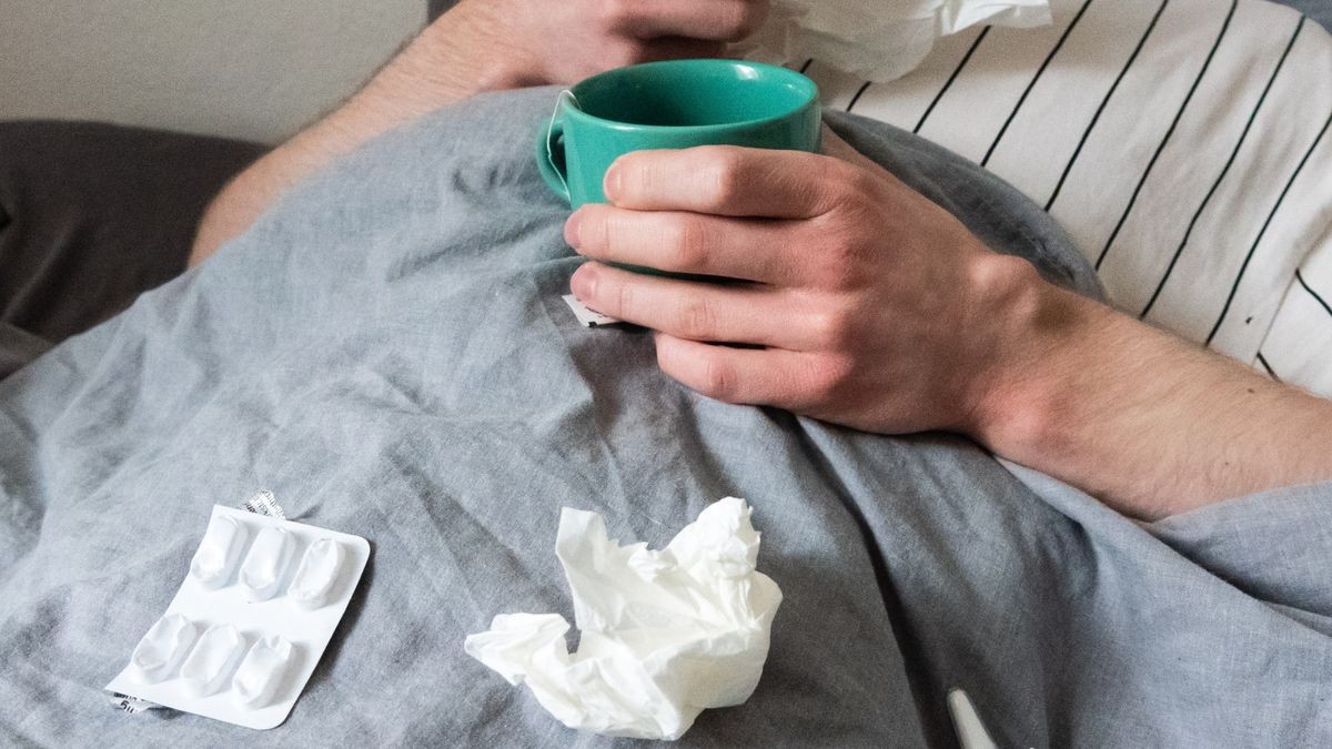 Ein Mann liegt mit Taschentüchern, Teetasse, Nasenspray und Tabletten im Bett.