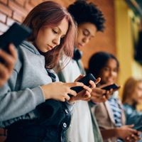 Schülerinnen und Schüler brauchen Auszeiten vom Handy, aber auch digitale Kompetenzen. Group of teenagers using mobile phones in hallway at high school.