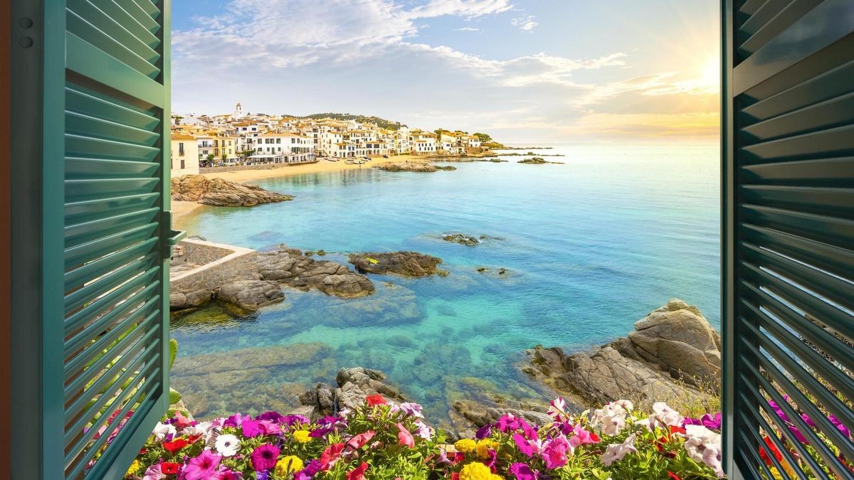View through an open window with shutters of the sandy beach, rocky coastline and whitewashed town of Calella de Palafrugell, Spain, on the Costa Brava coast as the sun sets.