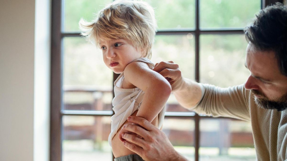Father with small sick unhappy son indoors at home, checking his back.
