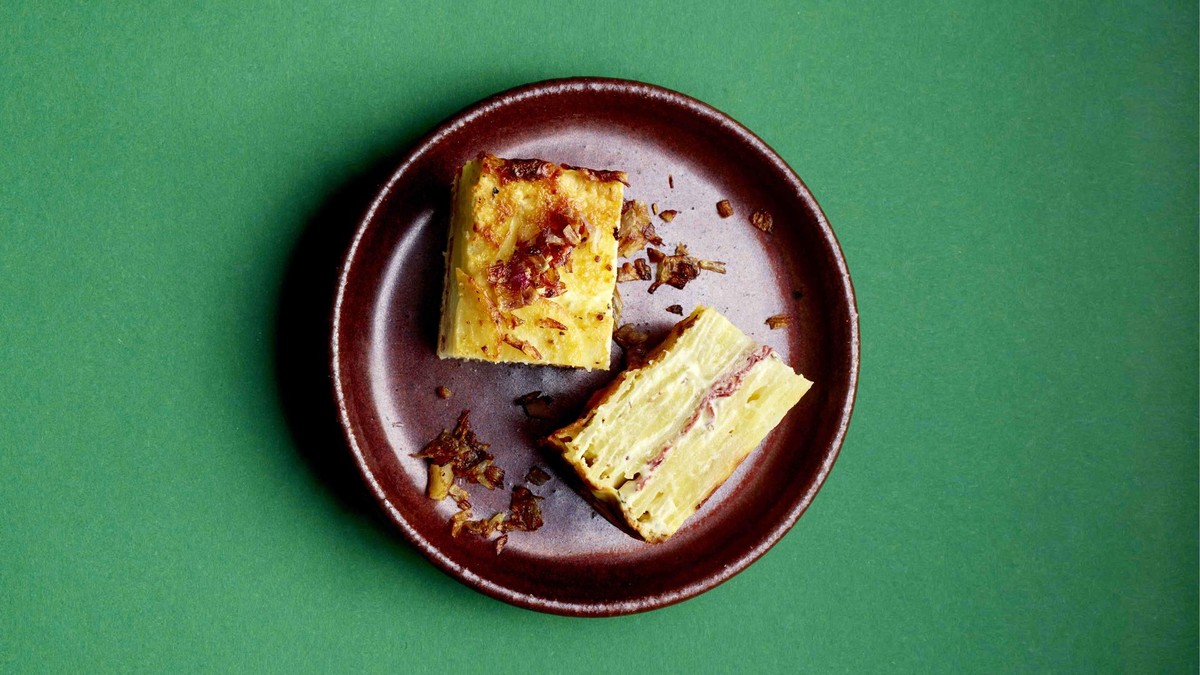 Ein Teller vor grünem Hintergrund mit zwei Stücken Kartoffelspeckgratin, dazu geröstete Schalotten